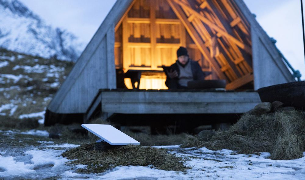 A Starlink Mini in the snow outside a lit A-frame hut at dusk