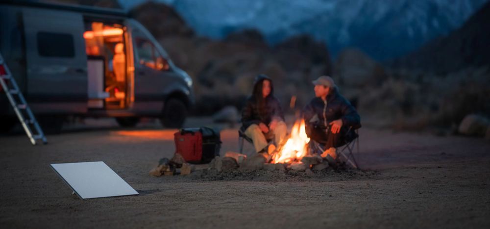 Two people at a campfire beside a van at dusk, a Starlink Mini on the ground nearby